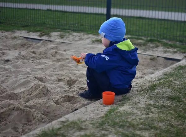 Küçük çocuk arka bahçedeki kum havuzunda bir kova ve kürekle oynuyor. Çocuk baharda dışarıda oyuncaklarla kum yemeği yapıyor..