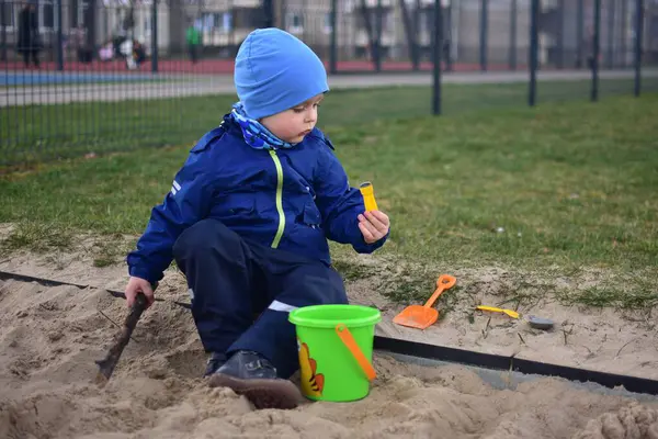Küçük çocuk arka bahçedeki kum havuzunda bir kova ve kürekle oynuyor. Çocuk baharda dışarıda oyuncaklarla kum yemeği yapıyor..