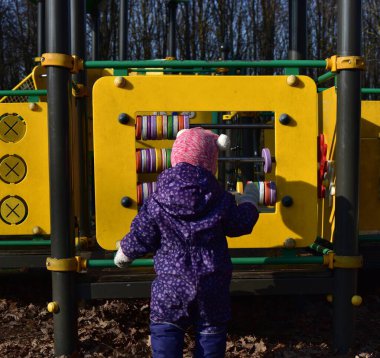 Yavru kız soğuk baharda şehir parkında çocuk parkında oyun oynuyor. Küçük kız kış güneşli bir günde halk parkında eğleniyor..