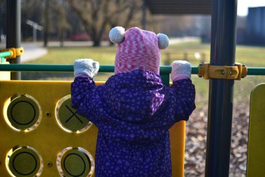 Yavru kız soğuk baharda şehir parkında çocuk parkında oyun oynuyor. Küçük kız kış güneşli bir günde halk parkında eğleniyor..