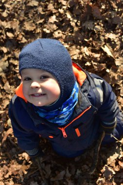 Küçük çocuk gülümsüyor, ağaçların dallarıyla oynuyor, ağaçların dallarıyla oynuyor ve güneşli bir sonbahar gününde, devrilmiş yaprakların arasında parkta oturuyor. Çocuk hayal gücünü kullanarak dışarıda eğleniyor..