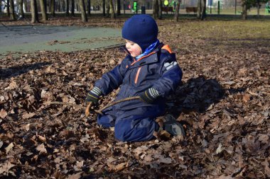 Küçük çocuk ağaç dallarıyla oynuyor ve güneşli bir sonbahar günü parkta düşen eski sonbahar yapraklarında oturuyor. Çocuk hayal gücünü kullanarak dışarıda eğleniyor..