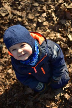 Küçük çocuk gülümsüyor, ağaçların dallarıyla oynuyor, ağaçların dallarıyla oynuyor ve güneşli bir sonbahar gününde, devrilmiş yaprakların arasında parkta oturuyor. Çocuk hayal gücünü kullanarak dışarıda eğleniyor..