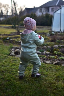 Naneli, yumuşak kabuklu bir beden giyen küçük kız baharda kırmızı ve siyah futbol topuyla arka bahçede oynuyor. Dışarıda çimenlerde yürüyen ve elinde top tutan küçük kız..