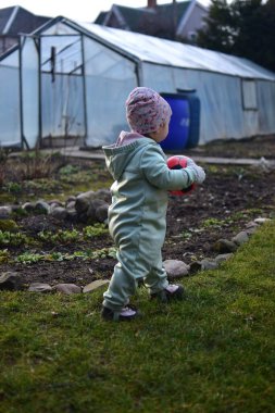 Naneli, yumuşak kabuklu bir beden giyen küçük kız baharda kırmızı ve siyah futbol topuyla arka bahçede oynuyor. Dışarıda çimenlerde yürüyen ve elinde top tutan küçük kız..