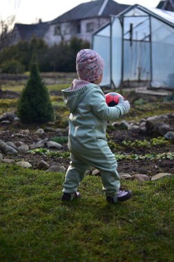 Naneli, yumuşak kabuklu bir beden giyen küçük kız baharda kırmızı ve siyah futbol topuyla arka bahçede oynuyor. Dışarıda çimenlerde yürüyen ve elinde top tutan küçük kız..