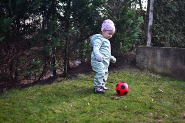 Naneli, yumuşak kabuklu bir beden giyen küçük kız baharda kırmızı ve siyah futbol topuyla arka bahçede oynuyor. Dışarıda çimenlerde yürüyen ve elinde top tutan küçük kız..