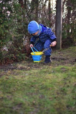 Mavi, yumuşak kabuklu mayolu çocuk baharda güneşli bir günde bahçedeki bir kovaya toprak kazıyor..