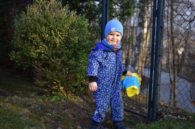 Mavi yumuşak kabuklu bebek elinde kova tutuyor ve güneşli bir bahar gününde arka bahçede toprakla oynuyor..