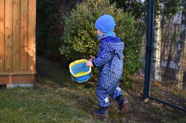 Mavi yumuşak kabuklu bebek elinde kova tutuyor ve güneşli bir bahar gününde arka bahçede toprakla oynuyor..