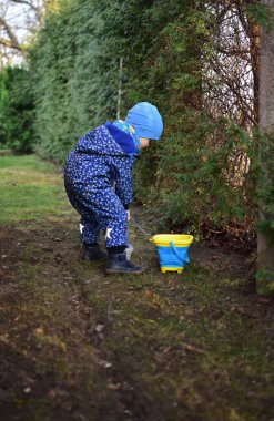 Mavi, yumuşak kabuklu mayolu çocuk baharda güneşli bir günde bahçedeki bir kovaya toprak kazıyor..