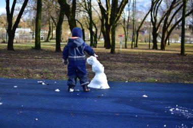 Küçük çocuk, ilkbaharda şehir parkındaki çocuk parkında neredeyse eriyen kardan adamın yanında duruyor. Çocuk ve üzgün görünümlü kardan adam kışın bitmesini bekliyor..