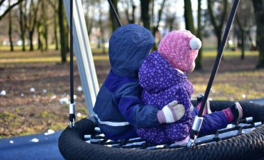 Erkek ve kız kardeş şehir parkında birlikte sallanıyorlar. İki kardeş arasındaki güçlü bağ kavramı. Erkek kardeşi küçük kız kardeşine sarılıyor. İki çocuk arkada oturuyor. Kardeşlerin sevgisi..