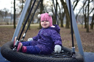Soğuk sonbaharda, şehir parkında çocuk parkında sallanan bir kız bebek. Küçük kız kışın halk parkında eğleniyor.