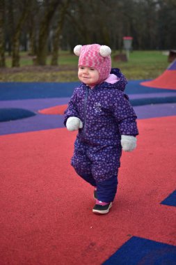 Yavru kız, soğuk baharda, şehir parkındaki renkli çocuk parkında gülümseyip oynuyor. Küçük kız kışın halk parkında eğleniyor..