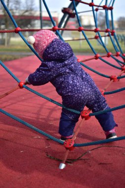Yavru kız gülümsüyor ve buz gibi baharda parktaki çocuk parkında ip tırmanışı yapıyor. Küçük kız kışın halk parkında eğleniyor..