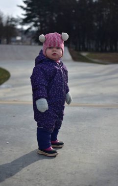 Yavru kız, soğuk baharda parktaki çocuk kaykay parkında duruyor. Küçük kız kışın boş kaykay parkında eğleniyor..