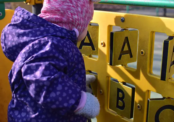 ABC ile oynayan küçük kız soğuk baharda şehir parkındaki çocuk parkında oyun oynuyor. Plastik dönen harfler ABC ekipmanları halka açık oyun parkına yakın görüntüde.