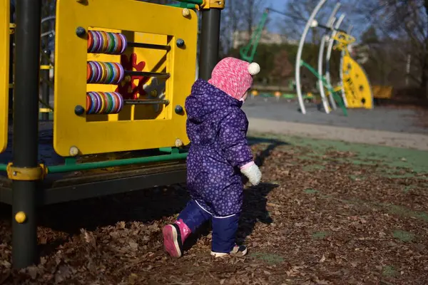 Yavru kız soğuk baharda şehir parkında çocuk parkında yürüyor ve oynuyor. Küçük kız kış güneşli bir günde halk parkında eğleniyor..