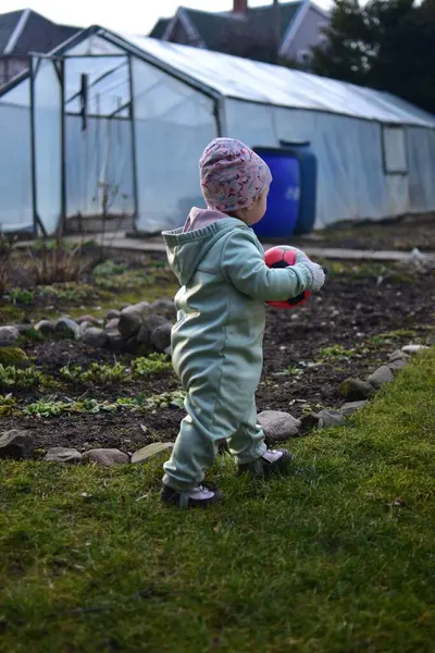 Naneli, yumuşak kabuklu bir beden giyen küçük kız baharda kırmızı ve siyah futbol topuyla arka bahçede oynuyor. Dışarıda çimenlerde yürüyen ve elinde top tutan küçük kız..