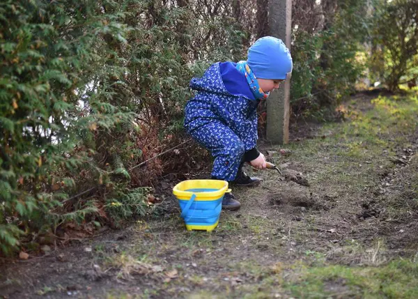 Mavi, yumuşak kabuklu mayolu çocuk baharda güneşli bir günde bahçedeki bir kovaya toprak kazıyor..