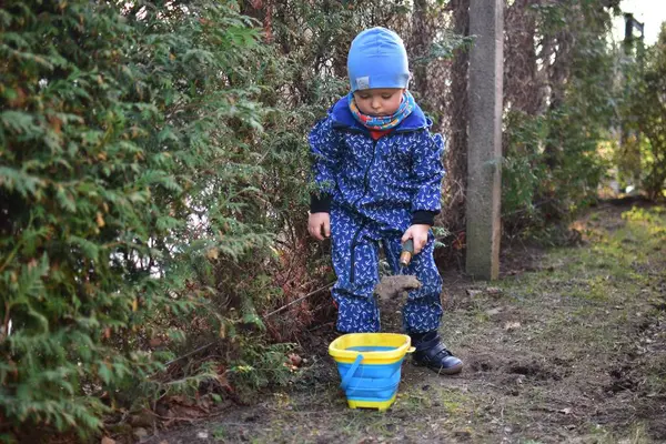 Mavi, yumuşak kabuklu mayolu çocuk baharda güneşli bir günde bahçedeki bir kovaya toprak kazıyor..