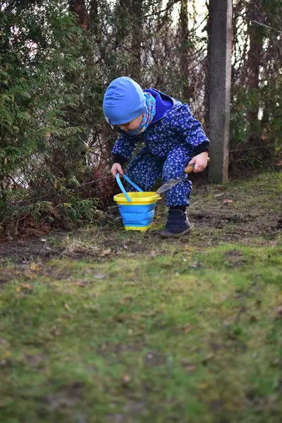 Mavi, yumuşak kabuklu mayolu çocuk baharda güneşli bir günde bahçedeki bir kovaya toprak kazıyor..