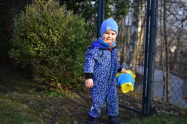Mavi yumuşak kabuklu bebek elinde kova tutuyor ve güneşli bir bahar gününde arka bahçede toprakla oynuyor..
