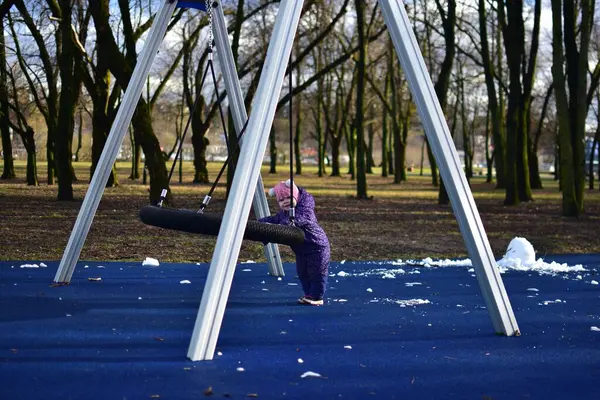 Yavru kız soğuk baharda salıncakların yanında duruyor ve parktaki çocuk parkında gülümsüyor. Küçük kız kışın halk parkında eğleniyor.