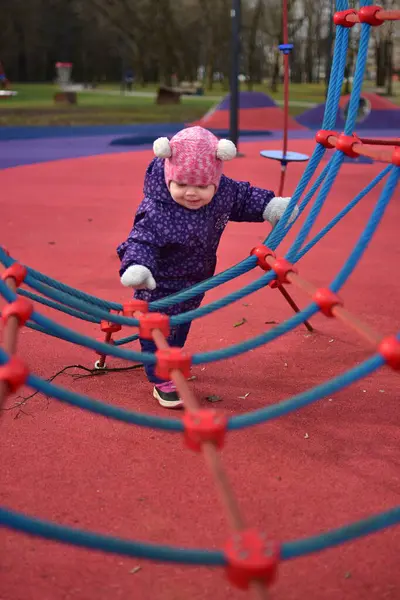 Yavru kız gülümsüyor ve buz gibi baharda parktaki çocuk parkında ip tırmanışı yapıyor. Küçük kız kışın halk parkında eğleniyor..