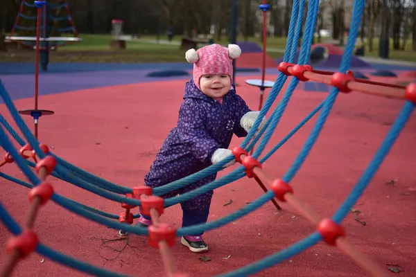 Yavru kız gülümsüyor ve buz gibi baharda parktaki çocuk parkında ip tırmanışı yapıyor. Küçük kız kışın halk parkında eğleniyor..