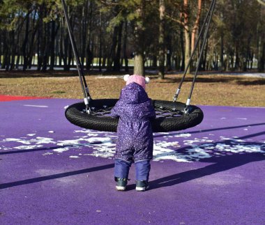Yavru kız soğuk sonbaharda şehir parkında çocuk parkında salıncakta sallanan sandalyeyle oynuyor. Küçük kız kışın halk parkında eğleniyor.