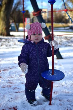 Bebek kız halat tutuyor ve soğuk havada parktaki karlı çocuk parkında oynuyor. Küçük kız kışın halk parkında eğleniyor.