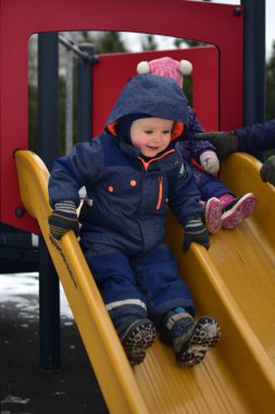Çocuk ve kız çocuk parkta çift kaydırak üzerinde oturuyor ve soğuk havada kaymaya hazırlanıyor. Erkek ve kız kardeş parkta birlikte oynuyorlar. Baba kızını tutuyor.