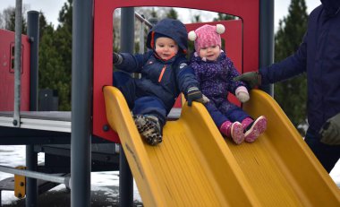 Çocuk ve kız çocuk parkta çift kaydırak üzerinde oturuyor ve soğuk havada kaymaya hazırlanıyor. Erkek ve kız kardeş parkta birlikte oynuyorlar. Baba kızını tutuyor.