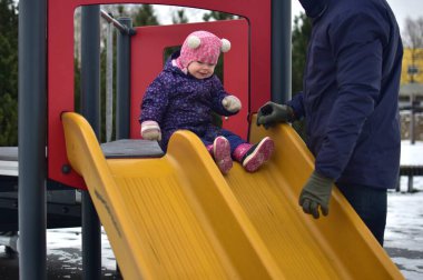 Küçük kız oyun parkında çift kaydırak üzerinde oturuyor ve soğuk havada kaymaya başlıyor. Baba kızını tutar ve aşağı kaymasına yardım eder..