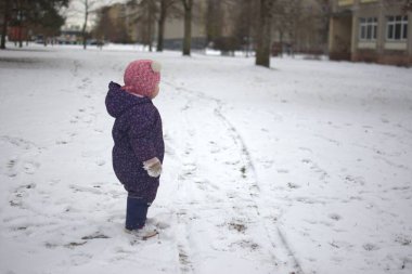 Yavru kız kar üzerinde duruyor ve soğuk kış aylarında ayak izlerine bakıyor. Küçük kız kışın dışarıda eğleniyor..