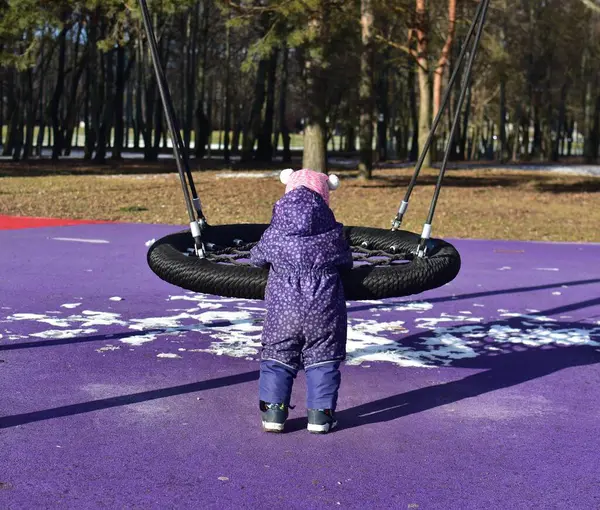 Yavru kız soğuk sonbaharda şehir parkında çocuk parkında salıncakta sallanan sandalyeyle oynuyor. Küçük kız kışın halk parkında eğleniyor.