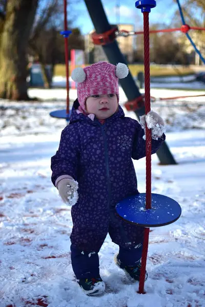 Bebek kız halat tutuyor ve soğuk havada parktaki karlı çocuk parkında oynuyor. Küçük kız kışın halk parkında eğleniyor.