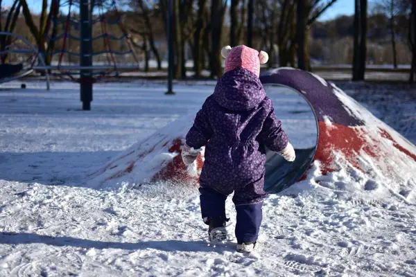 Yavru kız soğuk havalarda arka planda duruyor ve karlı çocuk parkında oyun oynuyor. Küçük kız kışın halk parkında eğleniyor.
