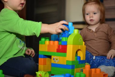 Küçük çocuk evde küçük kız kardeşiyle renkli bloklarla oynuyor. Küçük çocuk konsantre ve blok kale inşa ediyor gibi görünüyor..