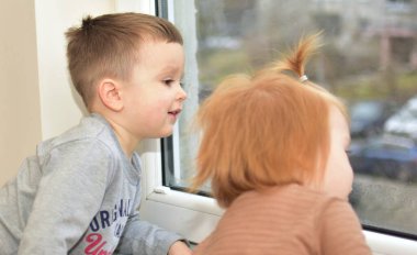 Küçük çocuk ve kız merakla pencereden bakıyor. Erkek ve kız kardeş sabırsız bir şekilde anne babalarını bekliyor..