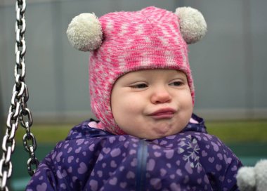 Bebek kızın portresi sert, çenesi kapalı soğuk sonbaharda şehir parkındaki çocuk parkında oynuyor. Küçük kız kışın halk parkında eğleniyor.