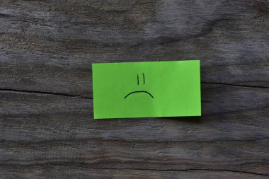 Ahşap kaplamalı arka planda yeşil yapışkan nottaki üzgün surat. Not kağıdına üzgün bir surat çiz..