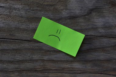 Ahşap kaplamalı arka planda yeşil yapışkan nottaki üzgün surat. Not kağıdına üzgün bir surat çiz..