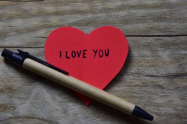Sevgililer günü için kırmızı kalp şeklindeki kağıtta, tahta bir arka planda, geri dönüşümlü kağıttan yapılmış sürdürülebilir kalemin yanında seni seviyorum mesajı. Sevgililer günü kartında 