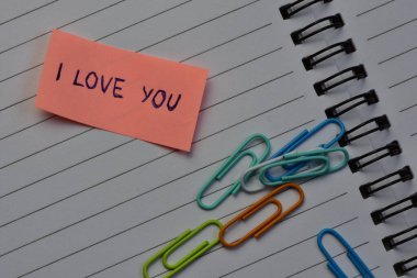 Sevgililer günü için pembe yapışkan notta, sarmal defter arkaplanındaki çeşitli renkli ataçların yanında seni seviyorum mesajı.