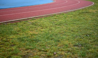 Sol tarafta mavi spor sahası olan kırmızı koşu pisti ve sağ tarafta yeşil çimenler, güneşli bir gün.