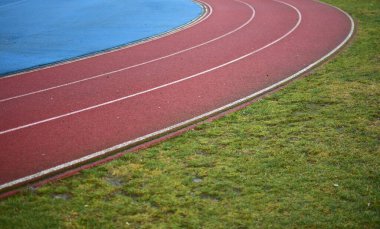 Sol tarafta mavi spor sahası olan kırmızı koşu pisti ve sağ tarafta yeşil çimenler, güneşli bir gün.