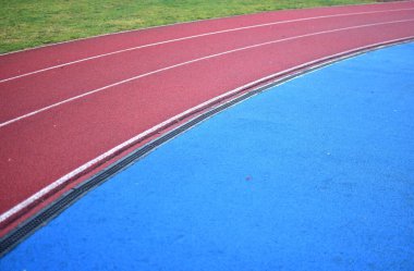 Kırmızı koşu parkuru sağ tarafta mavi spor sahası ve sol tarafta yeşil çimenler, güneşli bir gün.